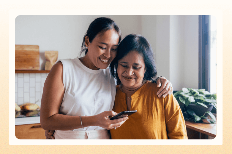 Adult daughter with arm around her mother looking at cell phone in hand
