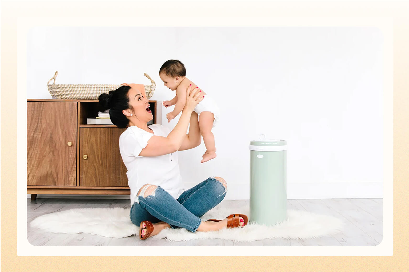 Woman sitting on floor next to a green diaper pail as she holds her baby up in the air with a big smile