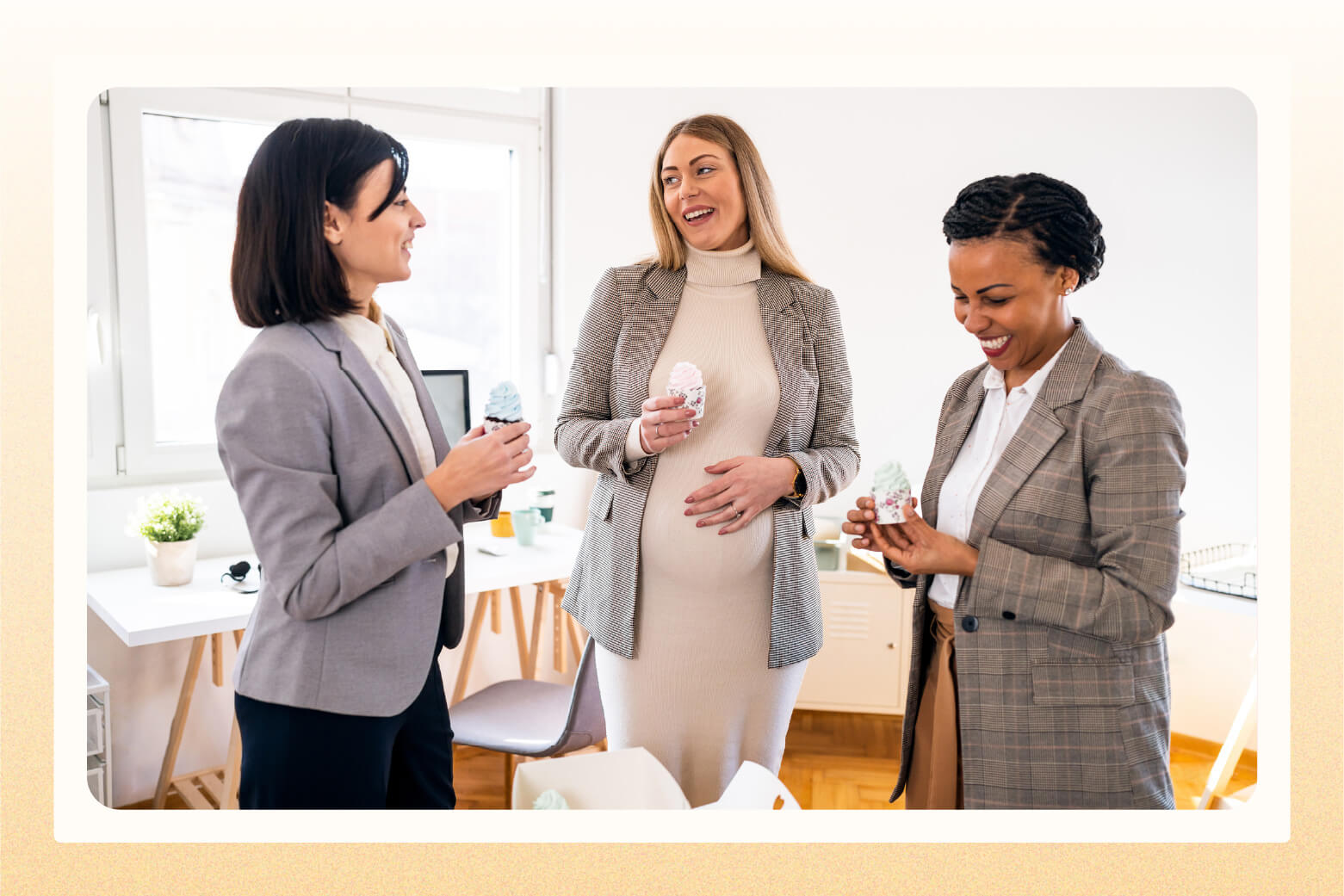 Three women in business attire stand in office holding cupcakes while talking to each other