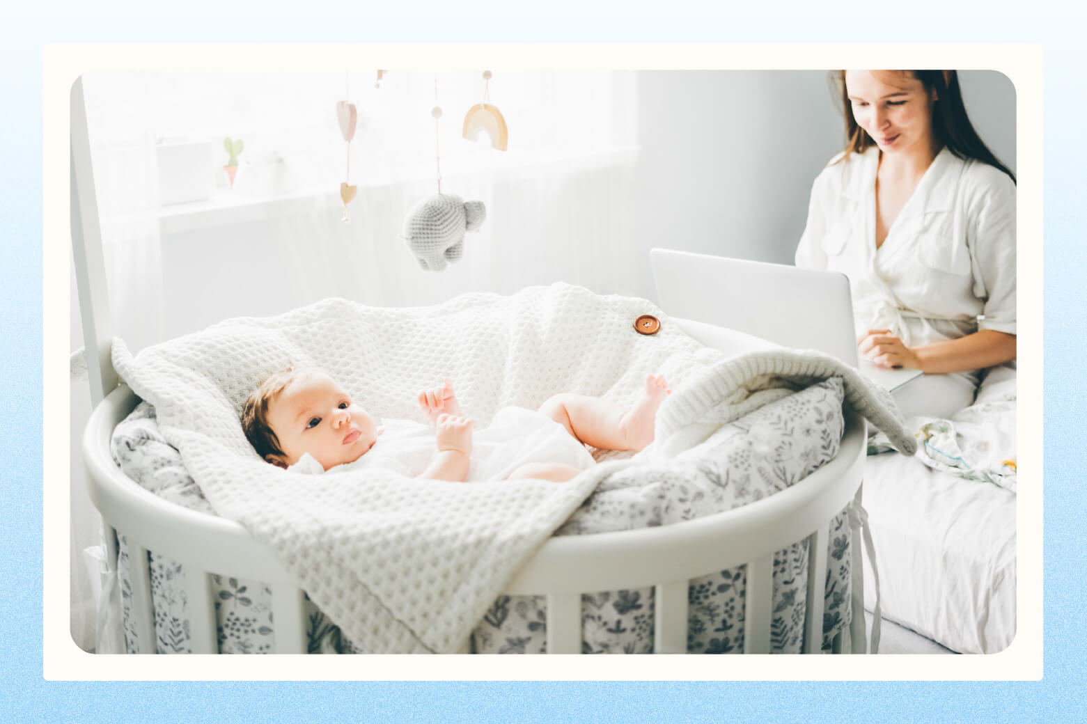 Woman on laptop sitting next to oval-shaped crib with baby laying inside of it on fluffy blankets