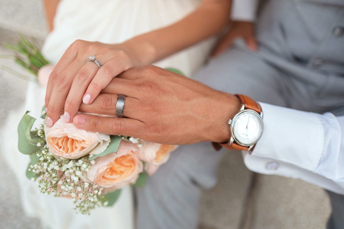 Engagement ring vs wedding ring: close-up of a couple in wedding attire holding hands and wearing an engagement ring and wedding ring