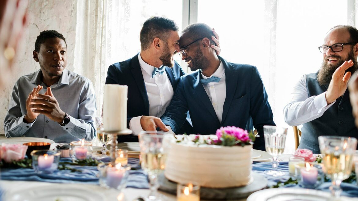 newlywed male couple sitting together and embracing one another at wedding reception table