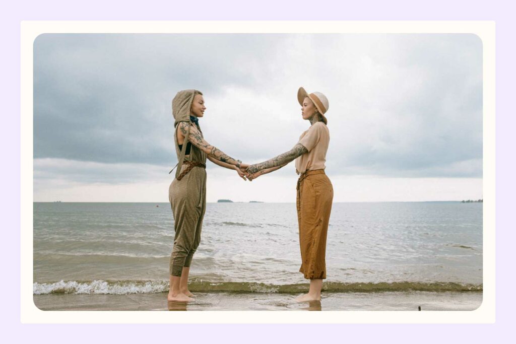 Lesbian couple standing facing each other on a beach, holding hands with arms outstretched