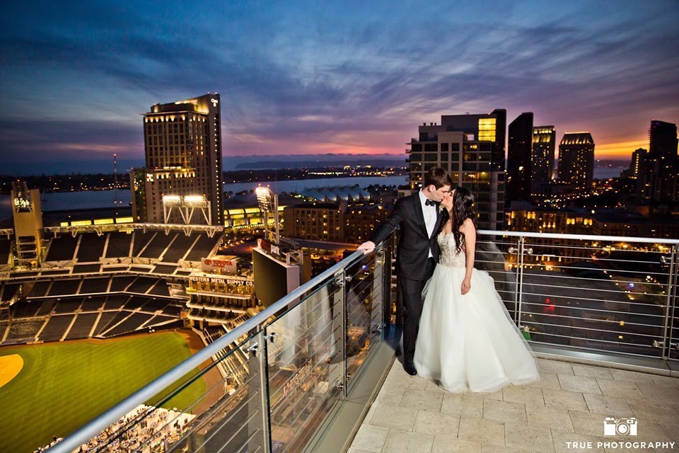 married couple kiss on the rooftop of diamond view tower in san diego with a view of the downtown skyline and baseball park