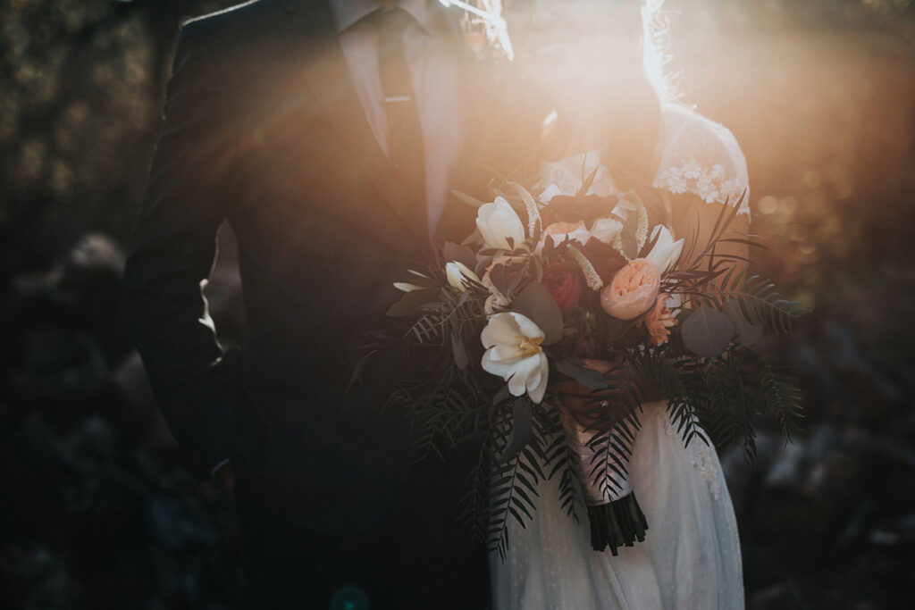 Couple in wedding attire standing in front of greenery and holding a bouquet of flowers with a glow of sunlight lighting the background
