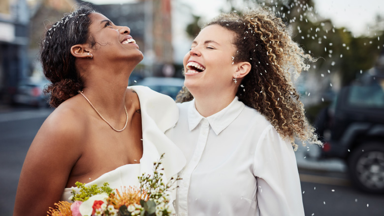 Two women in their wedding attire embracing with one arm around each other, heads thrown back with laughter