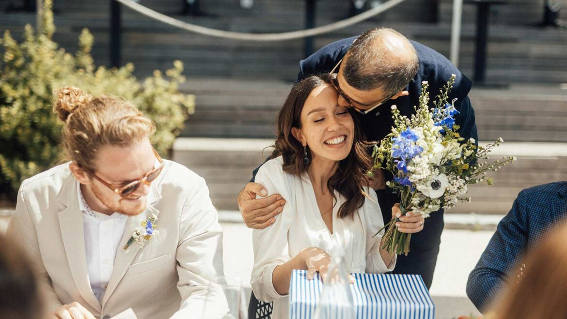 Woman about to open gift embraced by male family member with bouquet while male partner looks on with smile
