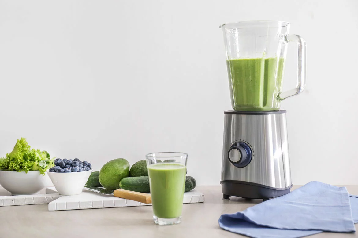 Blender on a kitchen countertop with a glass of green juice and bowls of fruits