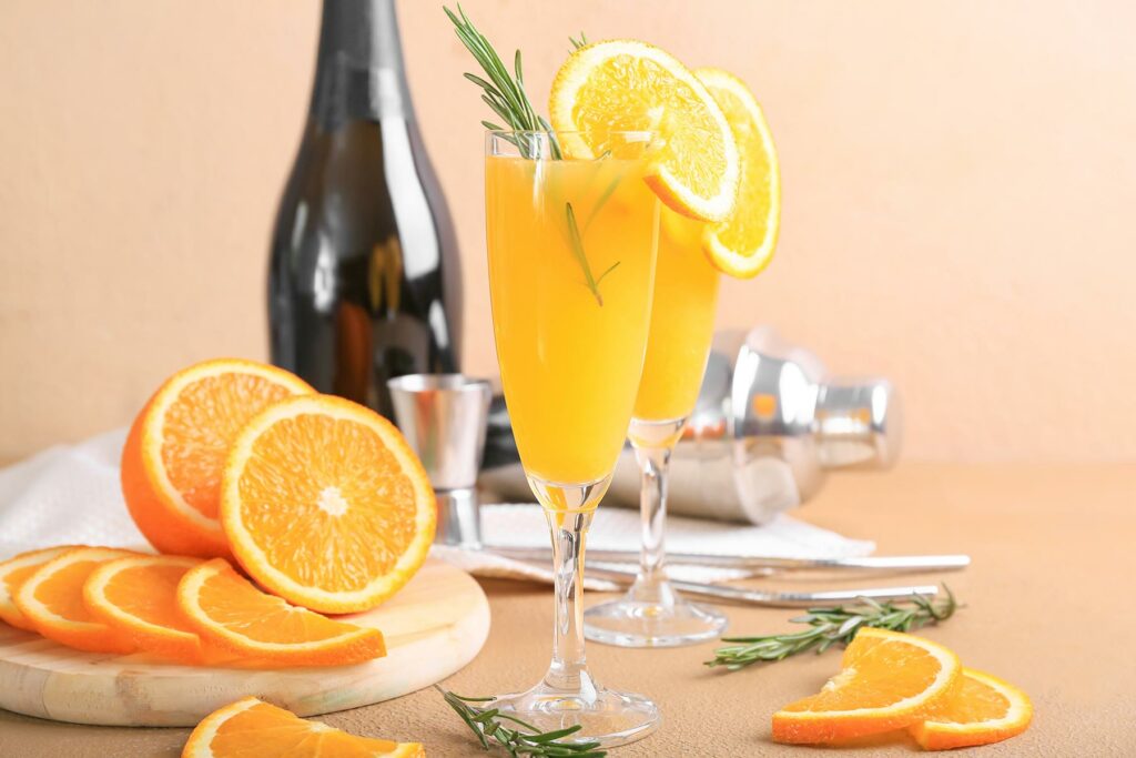Two mimosas garnished with orange slices in front of a bottle of champagne