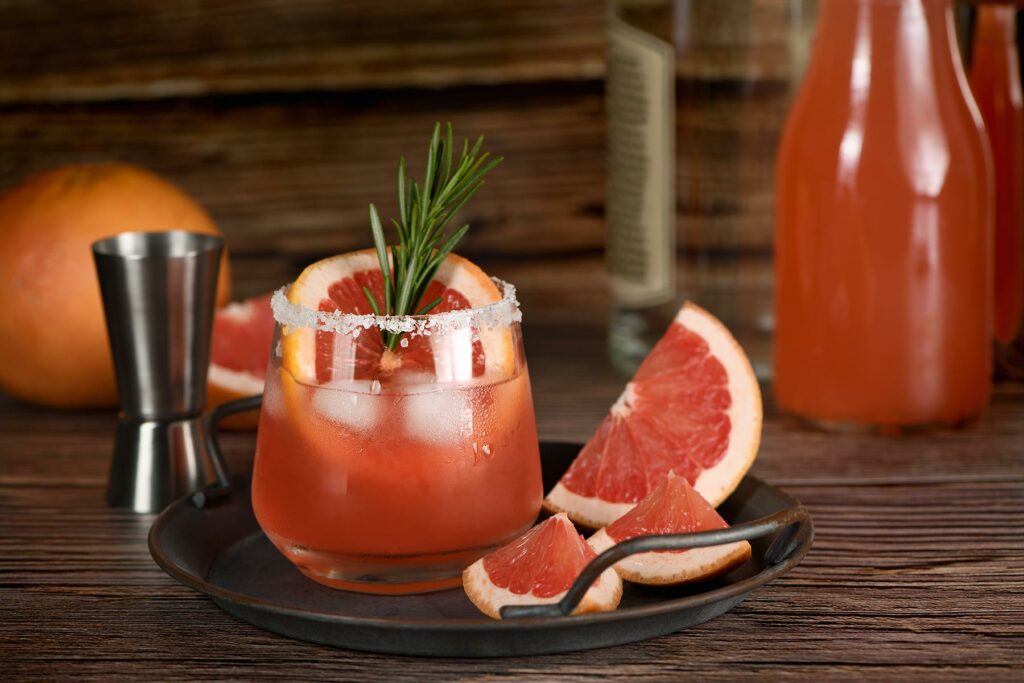 Paloma cocktail garnished with a sprig of rosemary on serving dish with wedges of grapefruit