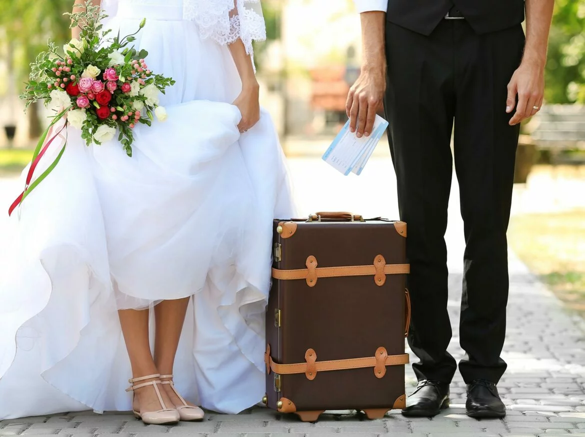 Couple in a wedding dress and suit standing next to a suitcase holding tickets for their honeymoon