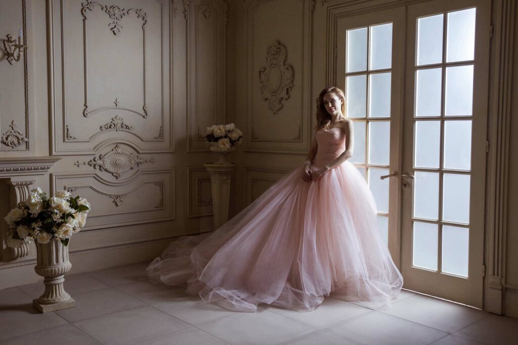 A woman wearing a blush pink wedding gown next to a window