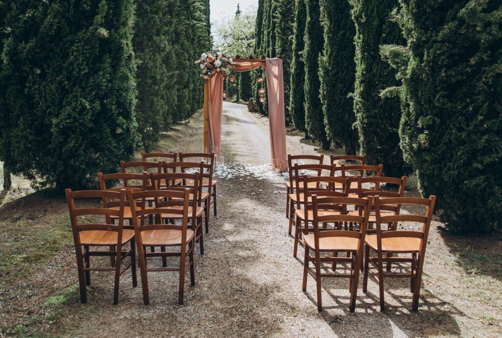 A backyard wedding ceremony is set up with eight chairs and a wedding arch in a grove of tall trees