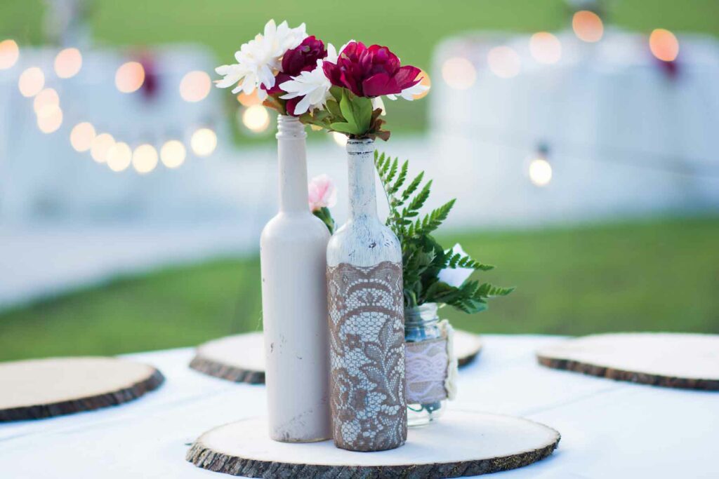 Spray painted bottles wrapped with lace and filled with dark red and white flower stems on a rustic tree slice