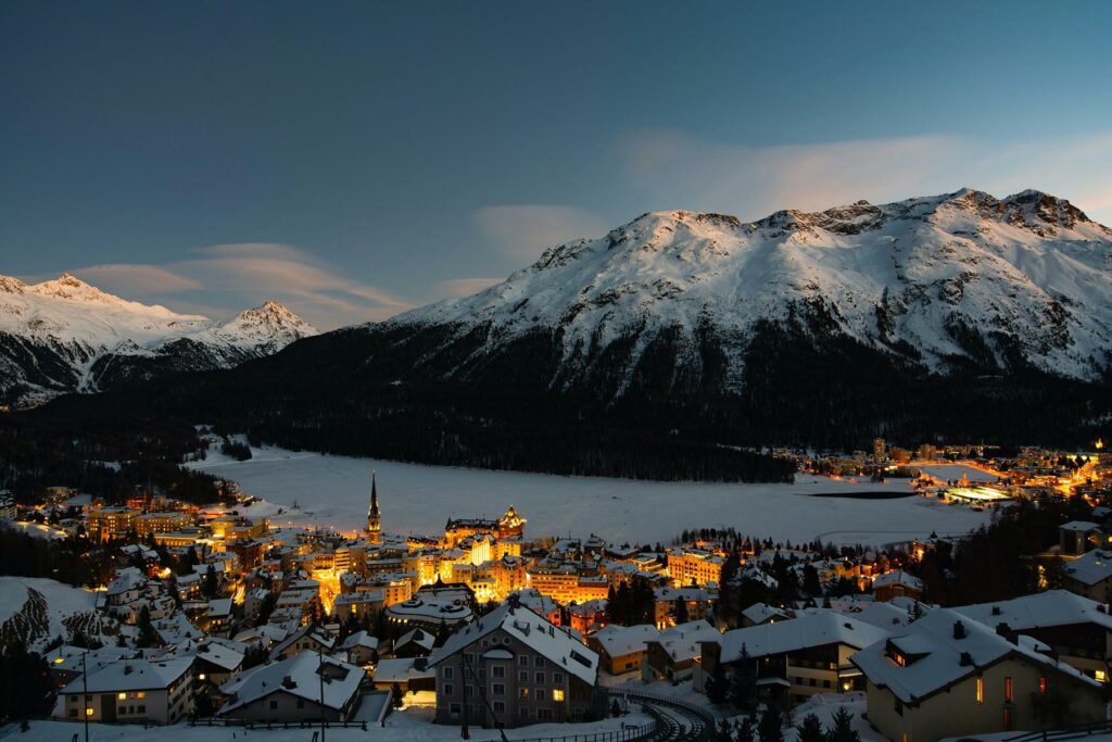 A view of mountains covered in snow and lit-up buildings from above in St. Moritz, Switzerland