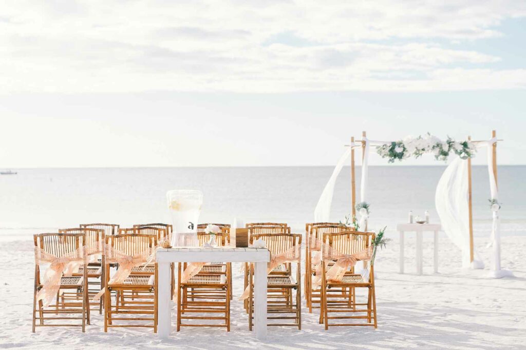 A beach wedding ceremony with wooden chairs and an arch with flowers