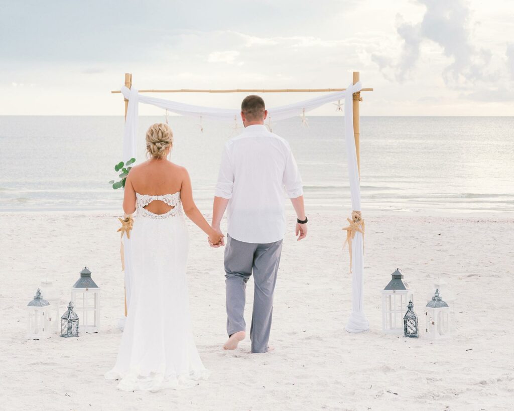 A couple at their beach wedding walking toward an arch draped with white fabric and surrounded by lanterns