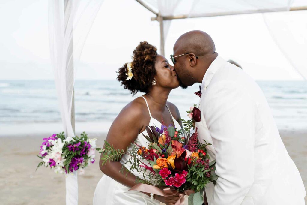 A couple kissing at their beach wedding ceremony under a canopy draped with white tulle