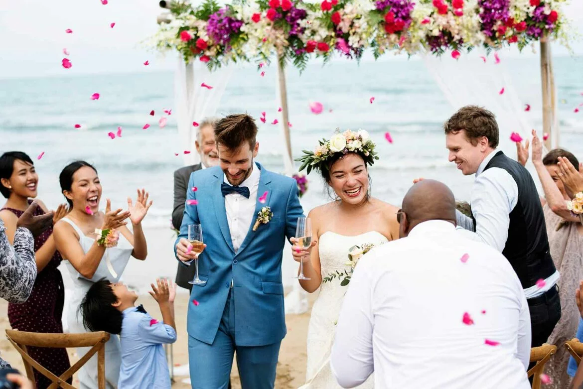 A couple at their beach wedding ceremony holding glasses of champagne and surrounded by loved ones