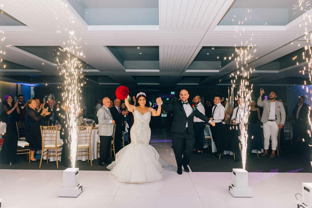 Guests clap and sparklers light up the room as a bride enters holding hands with the groom.