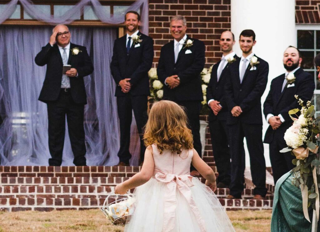 A flower girl walks down the aisle toward groomsmen and the groom during a wedding processional