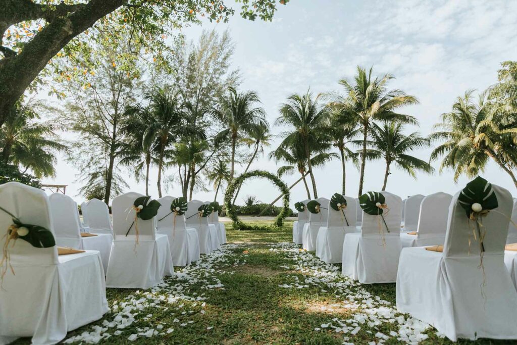 A destination wedding ceremony in a tropical setting with palm trees and a greenery archway at the end of an aisle