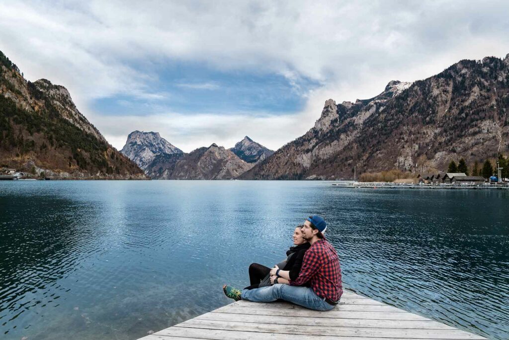 A couple on their honeymoon sitting on a dock looking up at mountains while surrounded by a lake