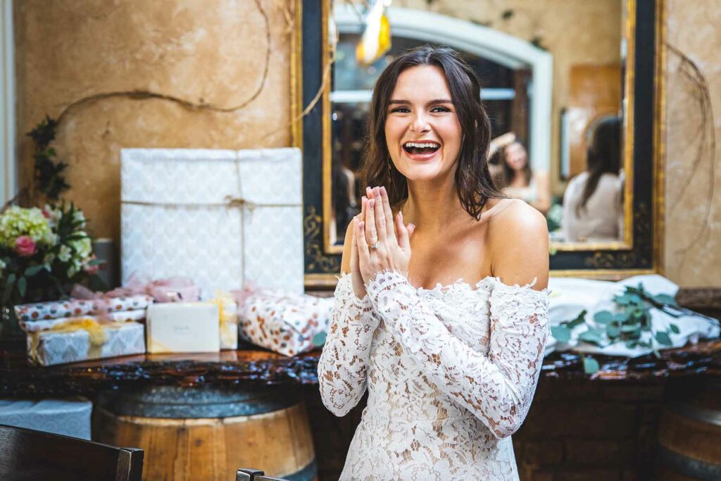 Woman in white lace dress smiles while standing in front of table filled with gifts