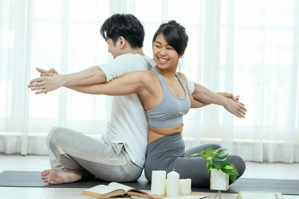 Couple dressed in yoga apparel and stretching arms and back while seated back-to-back and interlacing arms