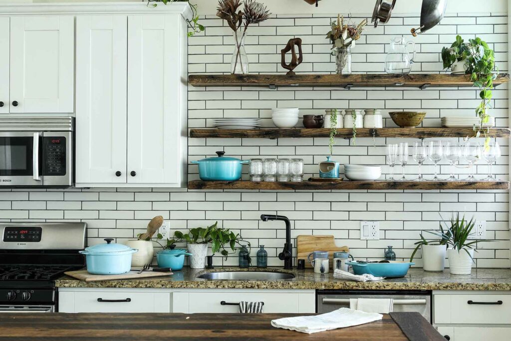 An open kitchen with wood shelves lined with wedding registry gifts like glassware, a dutch oven, plates and bowls, and a sink below