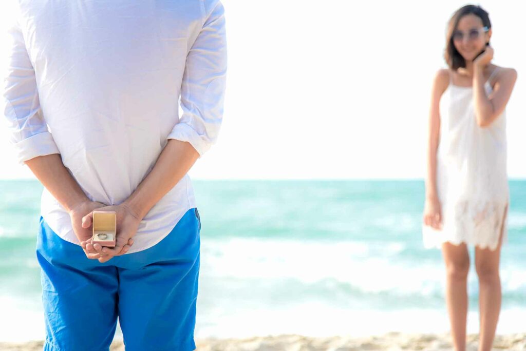 A man holding a ring behind his back during an outdoor proposal on the beach with a woman in white standing in the background