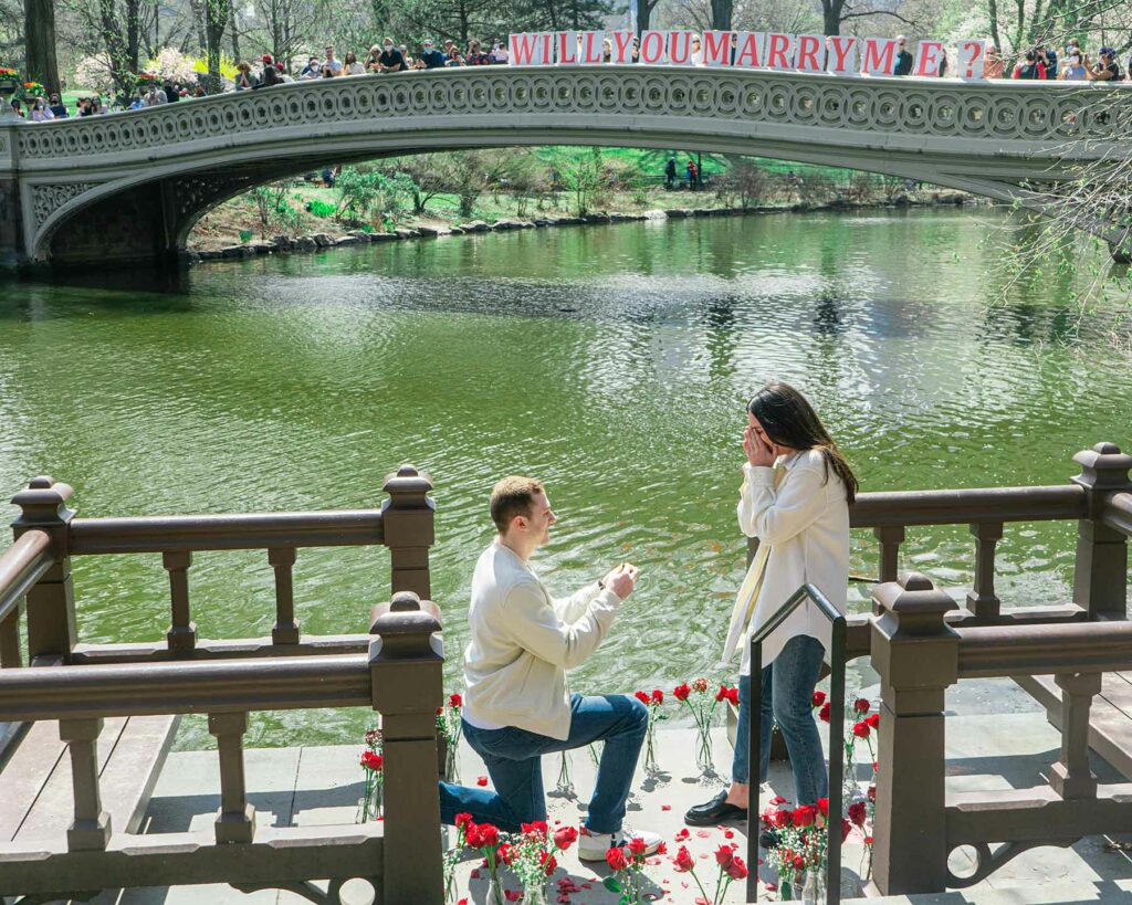 An outdoor proposal by a lake with a couple on a dock surrounded by flowers in faces and friends and family in the background on a bridge holding signs that read "will you marry me?"