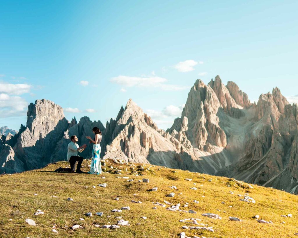 An outdoor proposal on a mountain with a person on one knee surrounded by rocky cliffs on a sunny day