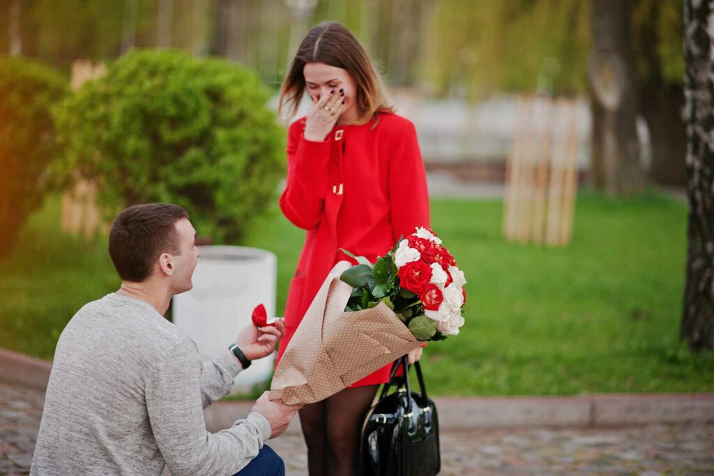 An outdoor proposal at a park with a man on one knee presenting a ring and a bouquet of flowers to a surprised woman