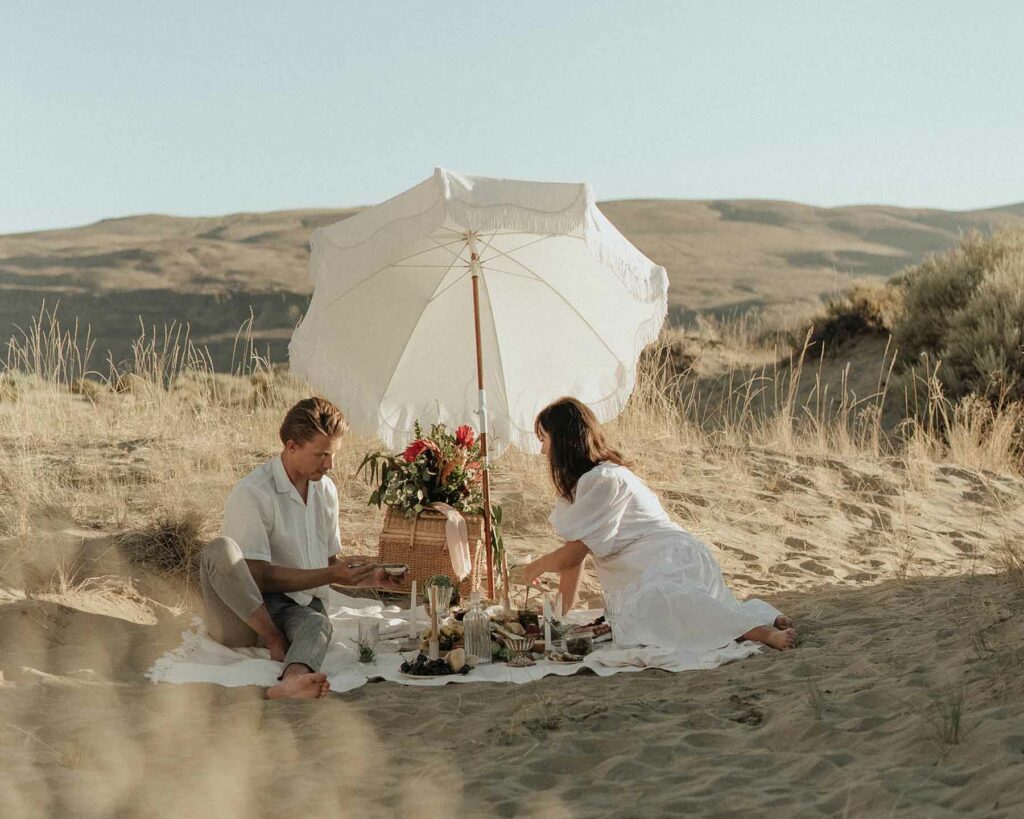 An outdoor proposal setup with a picnic on a beach and two people under an umbrella