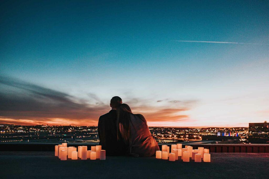 An outdoor proposal on a rooftop with two people sitting on a ledge surrounded by lit candles at twilight