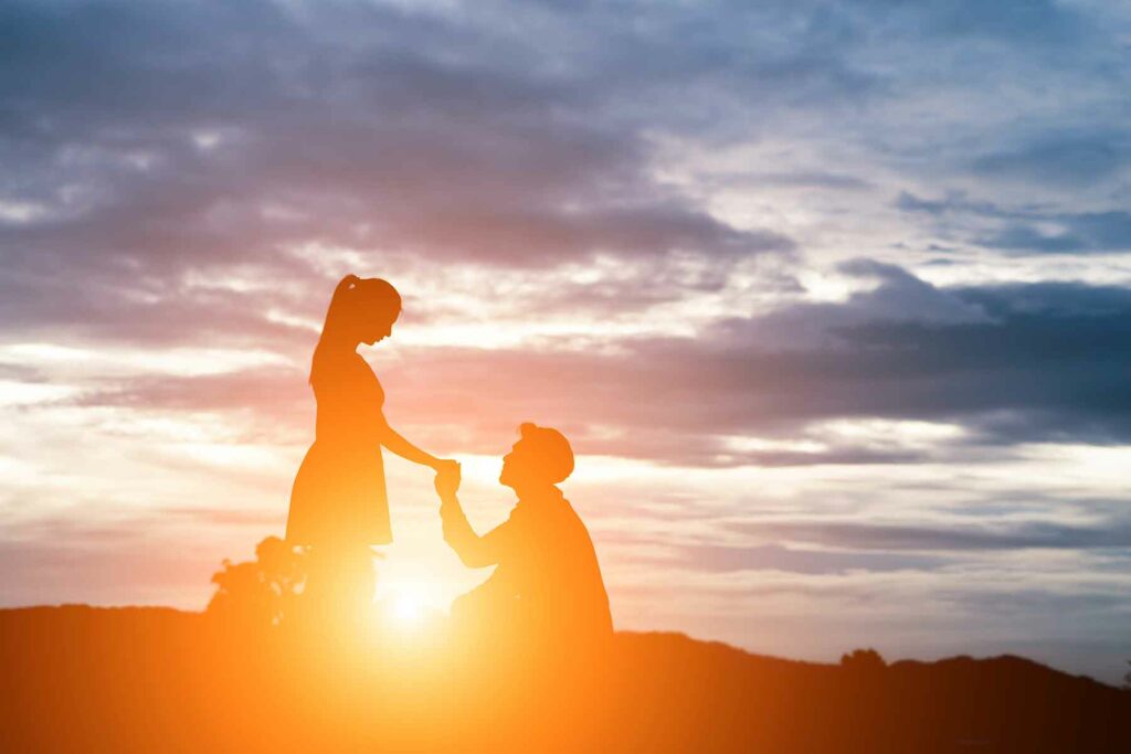 The silhouette of a person on one knee holding hands with a woman during an outdoor proposal at sunset