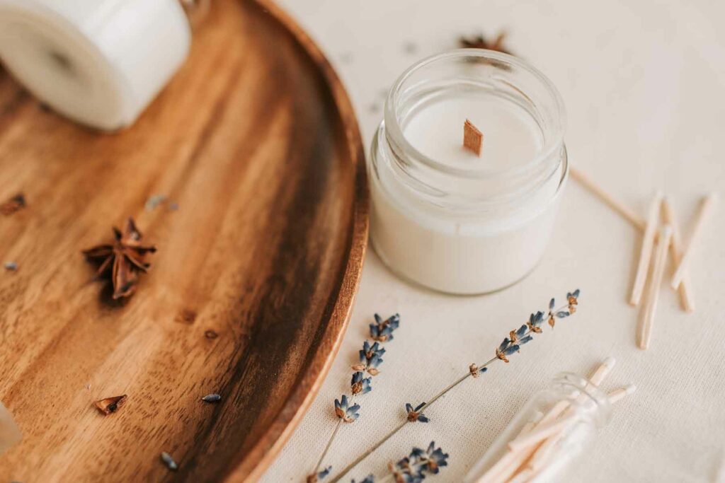 Close-up view of white candle with wooden wick in round glass jar next to matches, lavender and cloves