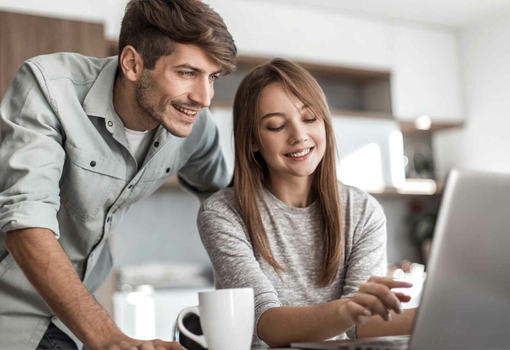 A couple in a kitchen looking at a laptop screen deciding where to register for their wedding