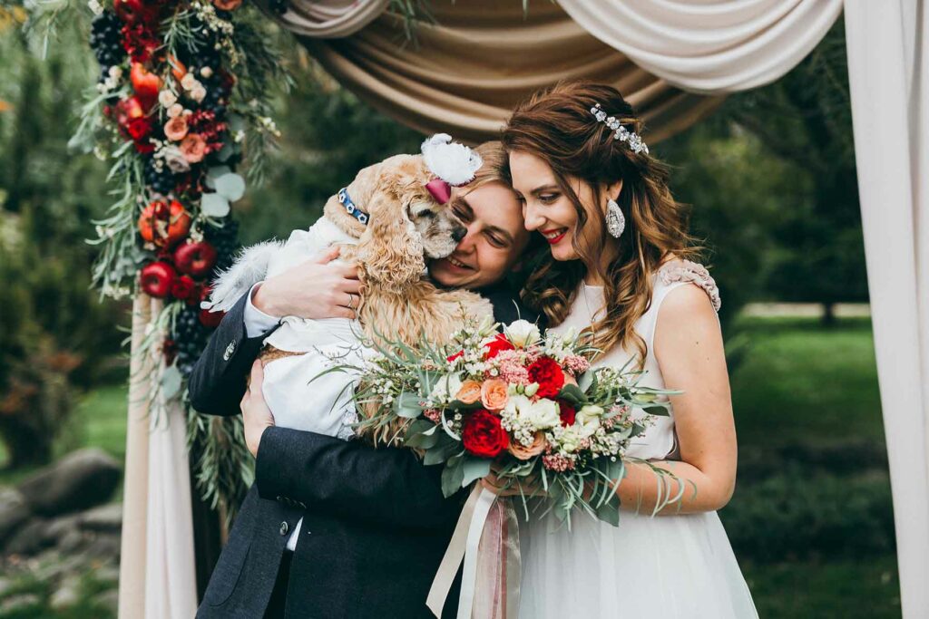 Fall wedding couple standing under an arch featuring apples and greenery and carrying a bouquet in fall colors of orange, gold and red