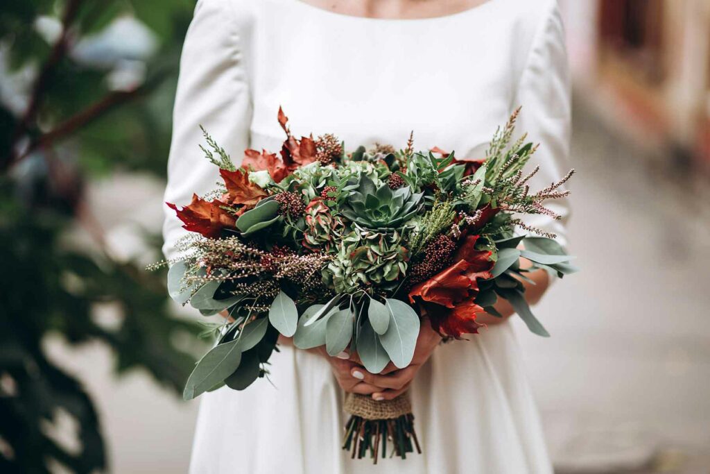 Bride holding a fall wedding bouquet with greenery, succulents and maple leaves wrapped in burlap