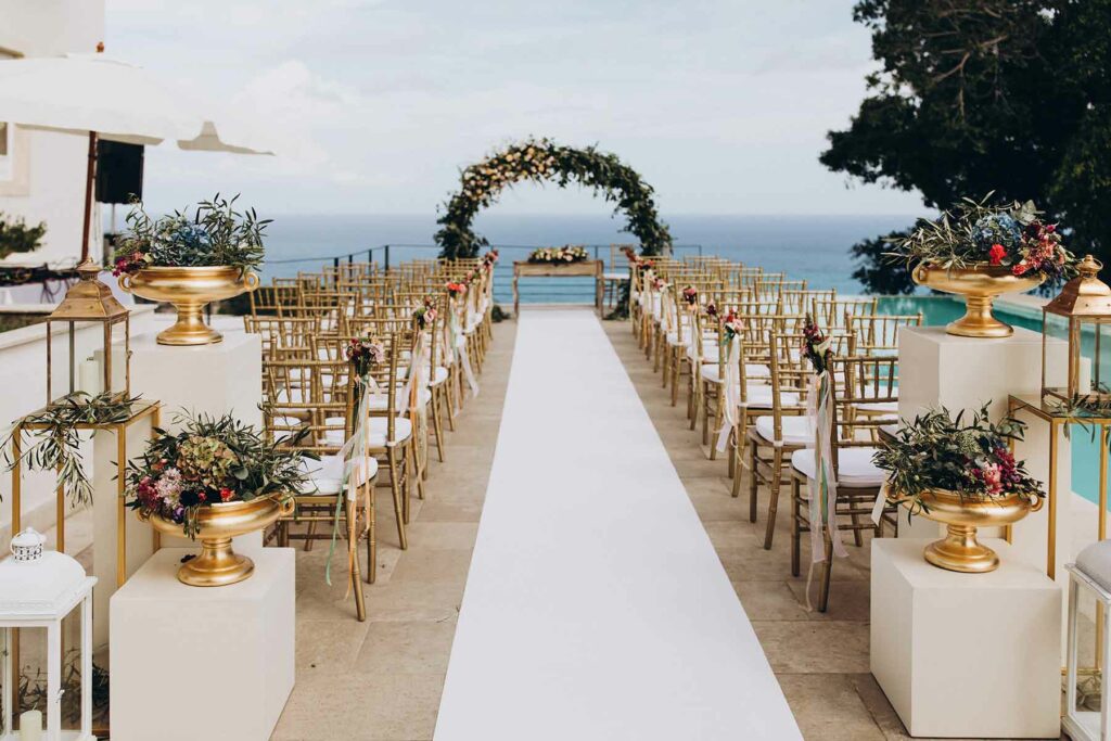Summer wedding with gold chairs, white aisle runner leading to a green arch surrounded by floral arrangements with warm undertones