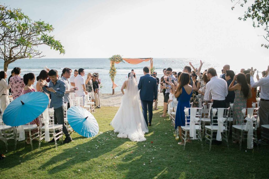 Summer wedding ceremony overlooking the beach with guests standing and parasols sitting next to the aisle