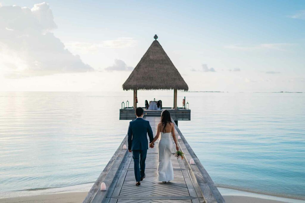 A couple at a destination wedding walking on an overwater bridge to a thatched roof hut