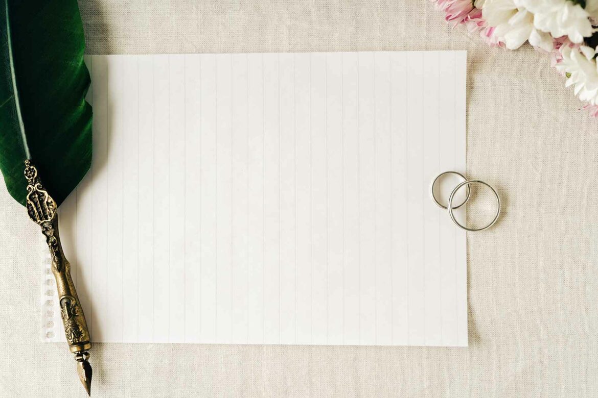 A feather calligraphy pen resting on a sheet of lined paper with two wedding bands and flowers on a table