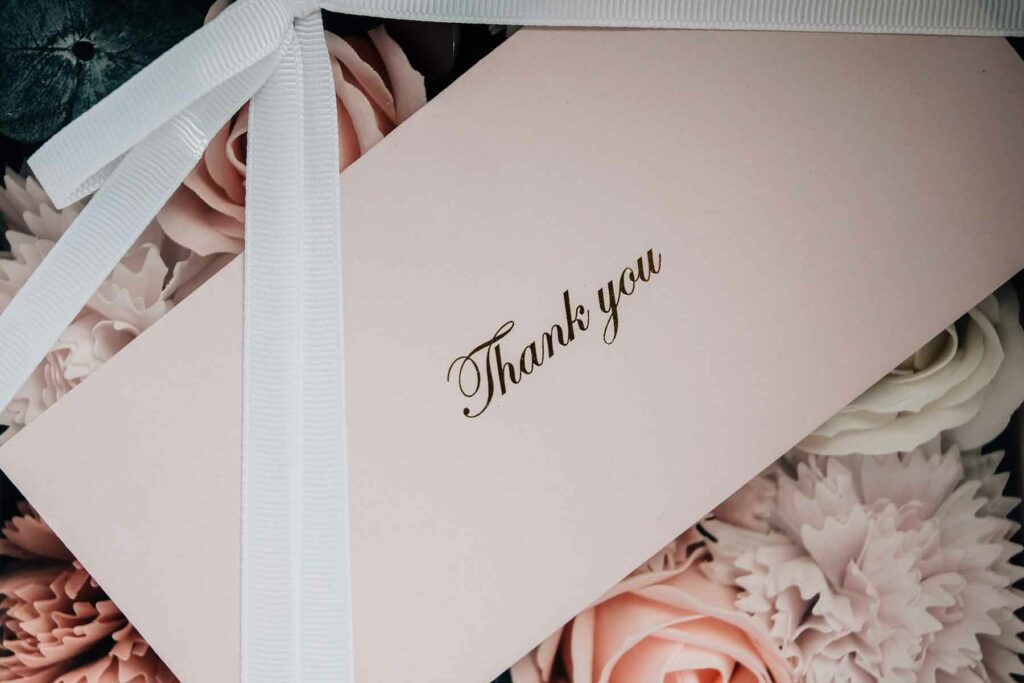 A folded card that reads "Thank you" in metallic script lying on top of a bed of wedding flowers.  There’s a white grosgrain ribbon tied into a bow laying over the thank you card. 