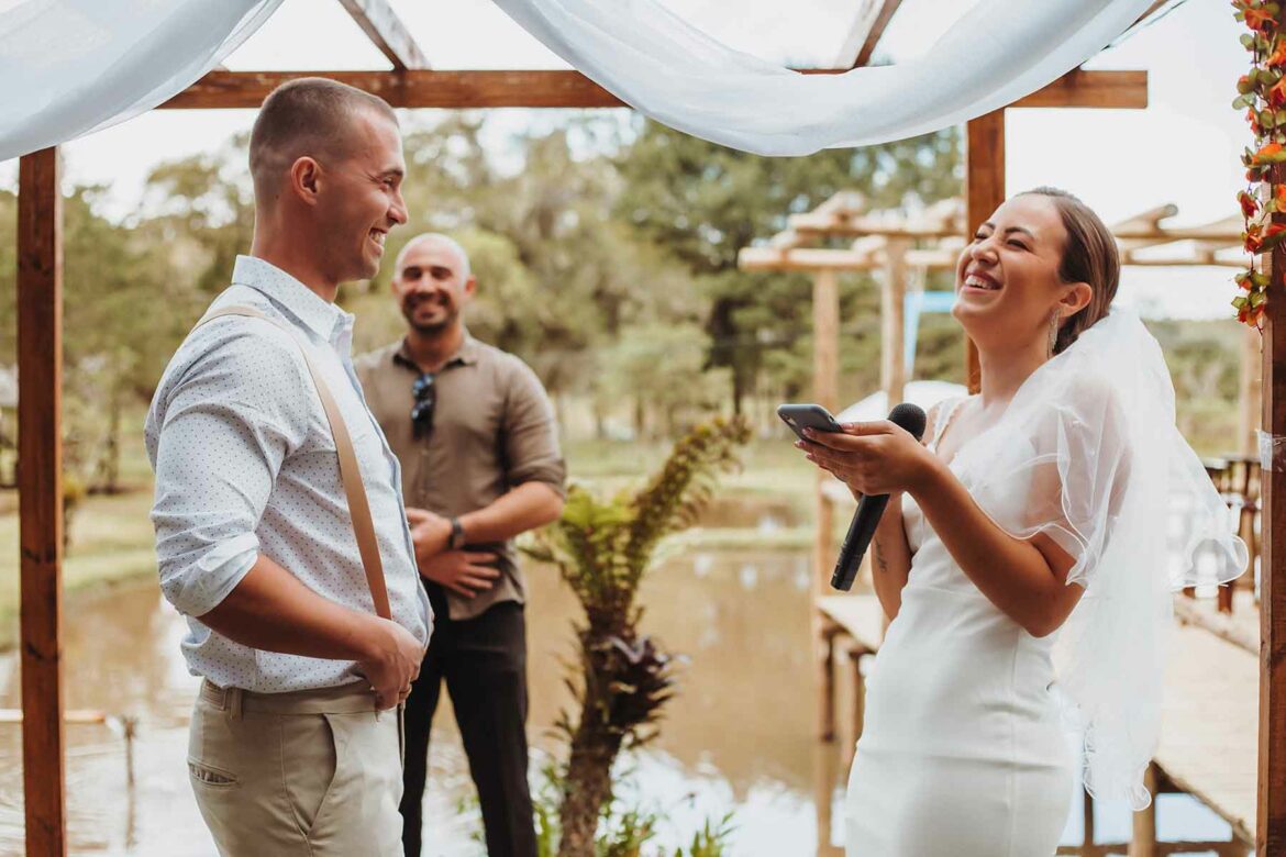 Micro-wedding photo of a couple by the water, laughing under a wooden canopy as they exchange vows