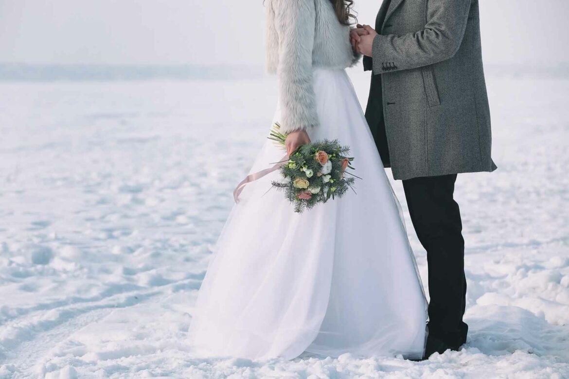 A woman in a faux fur jacket and wedding dress holds a bouquet, while clasping hands with a groom in a gray overcoat standing in snow at their winter wedding