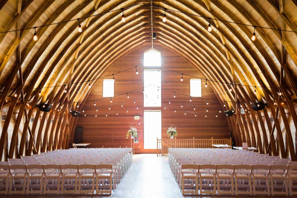 A ceremony venue with wood walls bathed in golden light. Rows of chairs are set up below strings of twinkling lights.