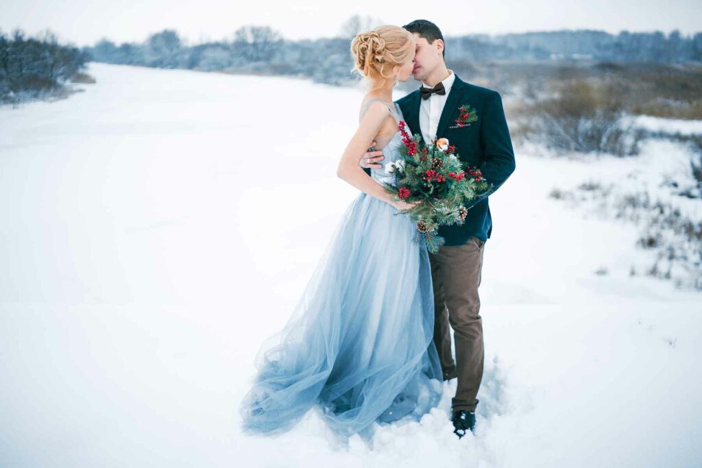 A couple kissing in the snow at their winter wedding. A woman in an icy blue wedding dress is holding a bouquet with pinecones and greenery. A man has a matching boutonniere